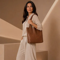Mulher de look claro usando a bolsa de ombro grande marrom moka, destacando o caimento macio do couro e o vão de 21cm.